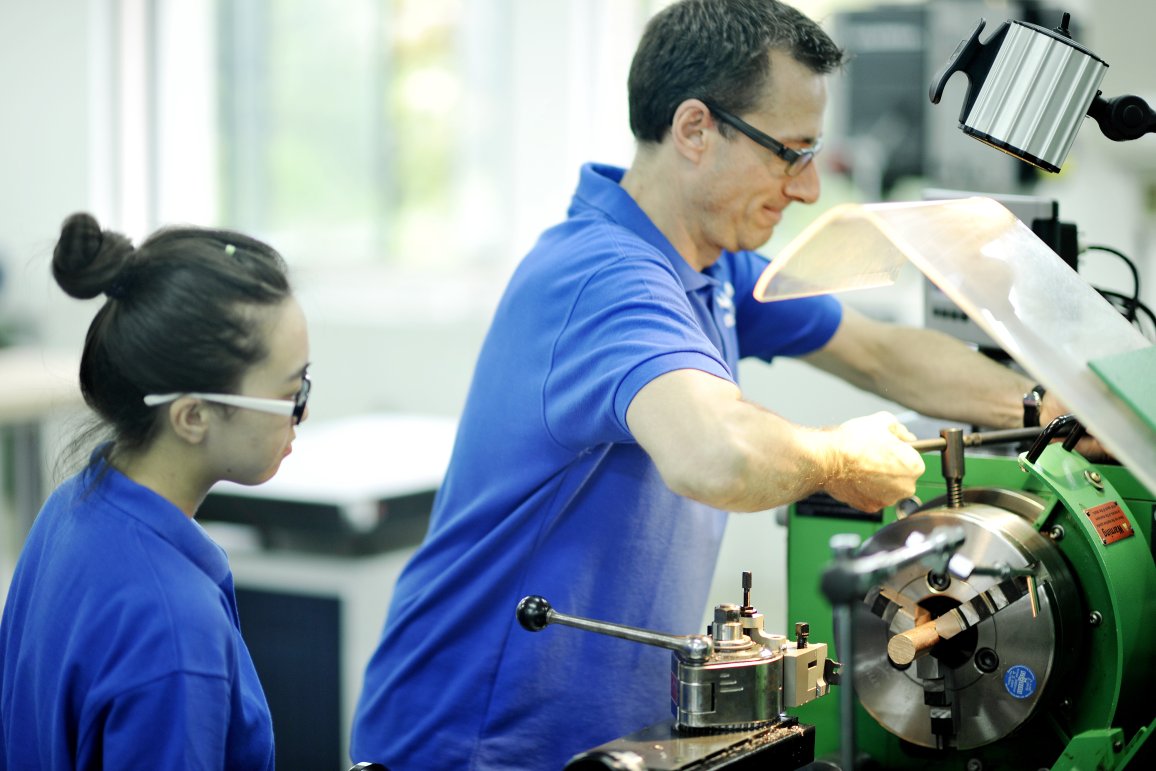 Juan Nitzl working at a machine with an apprentice watching him, shot 2014. 