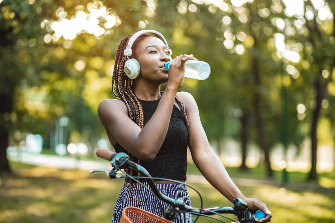 A black woman drinks from a PET bottle while standing with her bicycle.