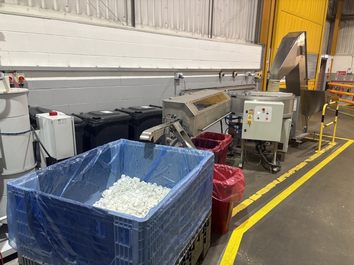 Blue bin filled with white plastic caps beside ALPLA packaging machinery in a clean factory.