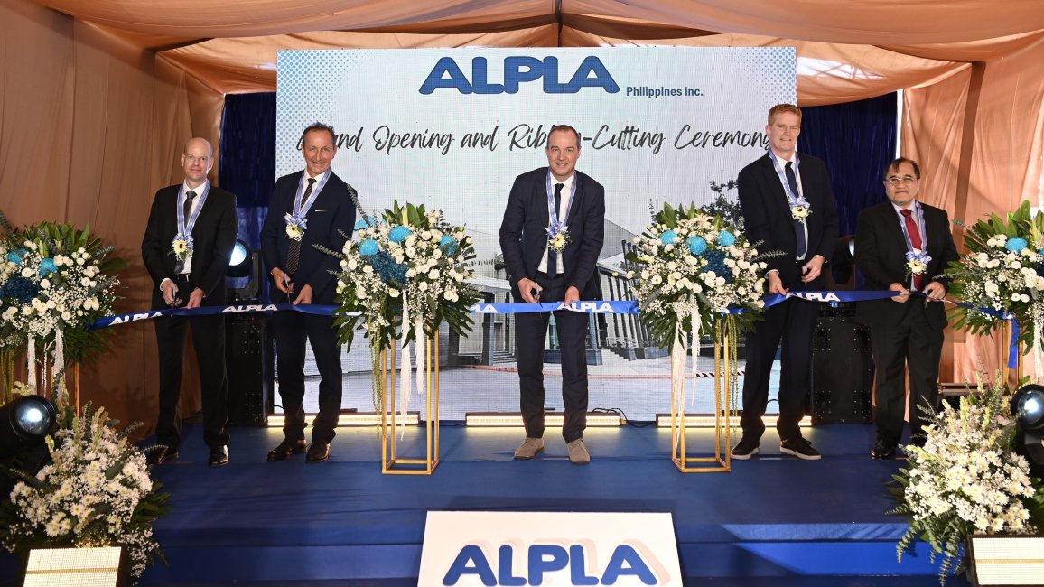 Executives cutting ribbon at ALPLA grand opening event with floral arrangements on stage.