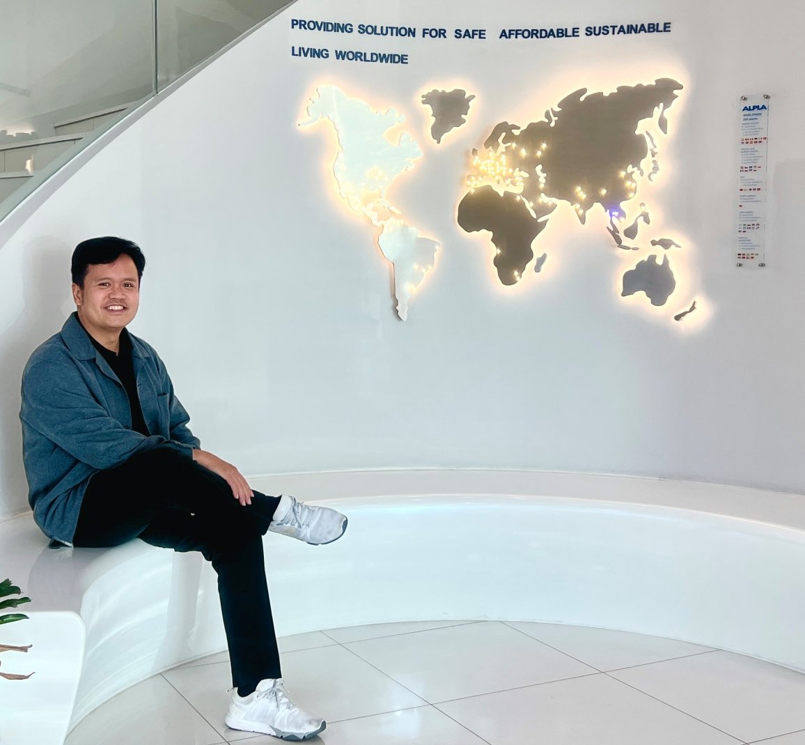 Ein lächelnder Mann sitzt auf einer weißen gebogenen Bank neben einer beleuchteten Weltkarte in einem modernen ALPLA-Büro