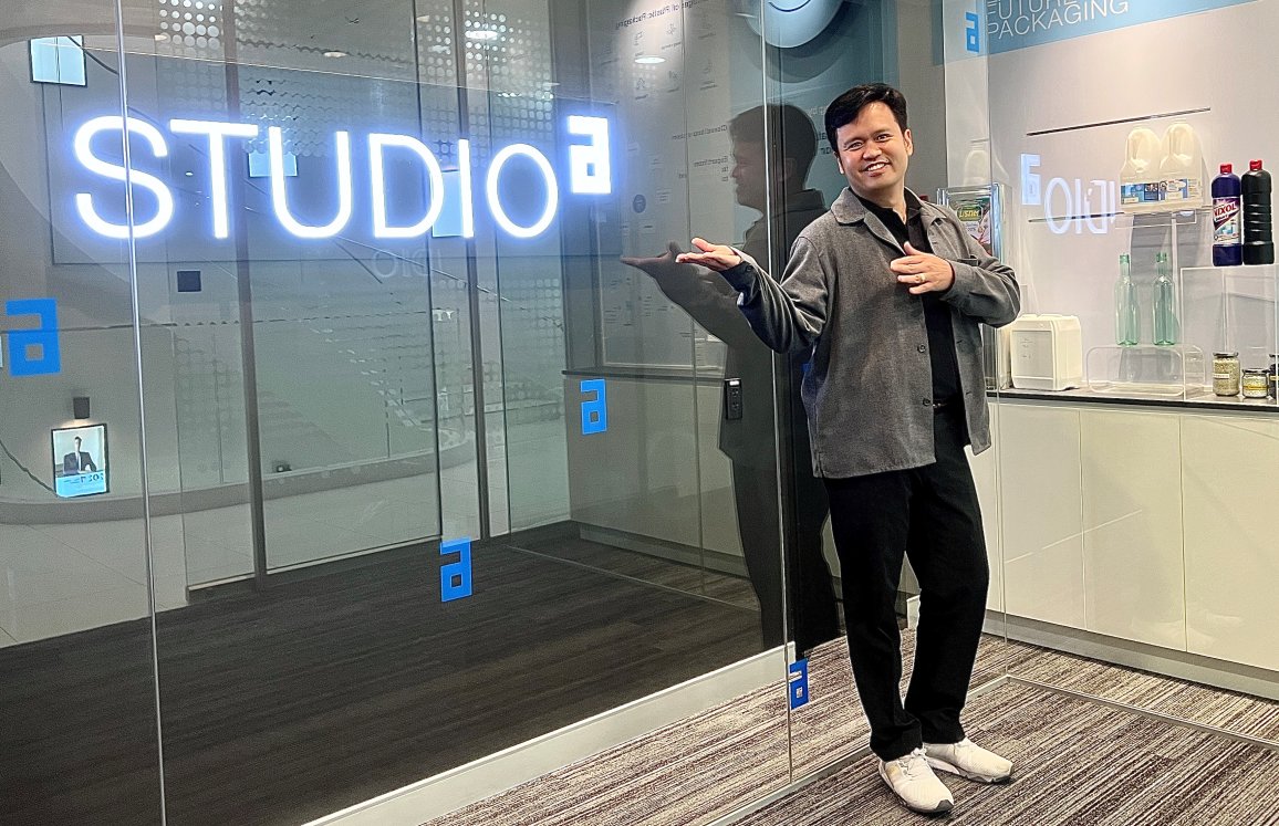 Lächelnde Person, die an einer Glaswand mit dem Schild "STUDIO a" steht, daneben eine ALPLA-Verpackung.
