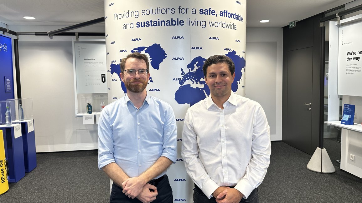 Two men in business attire standing in front of an ALPLA world map display.