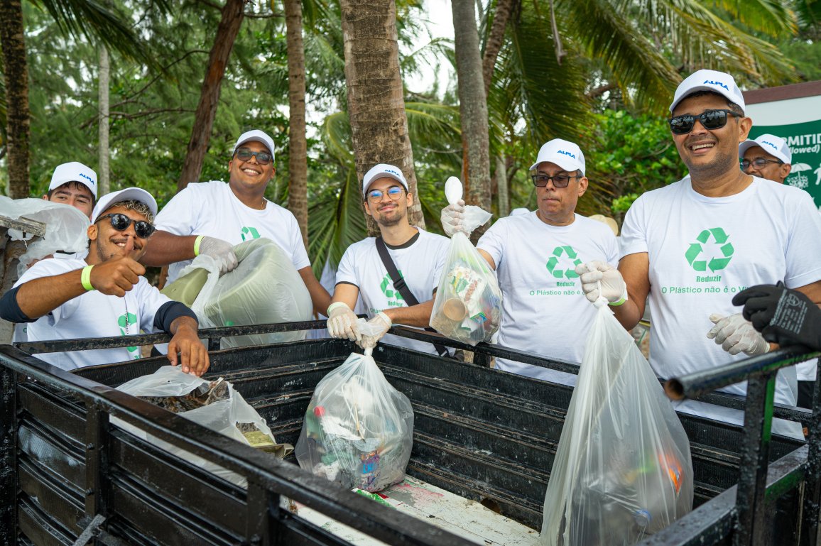 Teammitglieder in einheitlichen T-Shirts, die lächelnd im Freien Plastikmüll zum Recycling einsammeln.
