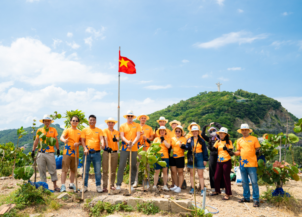 Gruppe von Menschen in orangefarbenen T-Shirts und Hüten pflanzt Bäume in Vietnam mit Christusstatue im Hintergrund.
