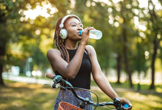A black woman drinks from a PET bottle while standing with her bicycle.