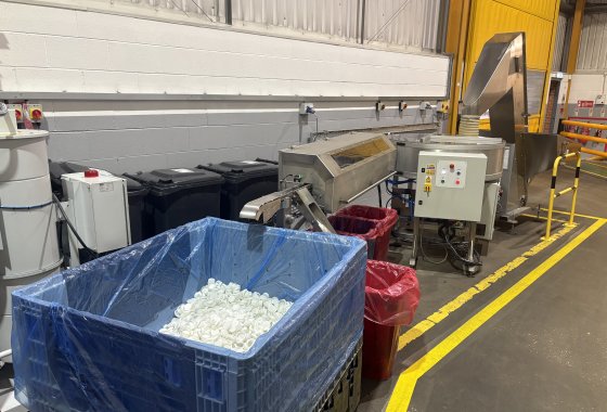 Blue bin filled with white plastic caps beside ALPLA packaging machinery in a clean factory.