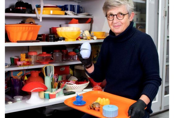 Eine Person mit kurzen grauen Haaren hält ein orangefarbenes Tablett mit Plastikverpackungen vor einem Verkaufsregal.