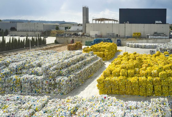 Bales of sorted plastic bottles stacked outside an ALPLA recycling facility under a cloudy sky.