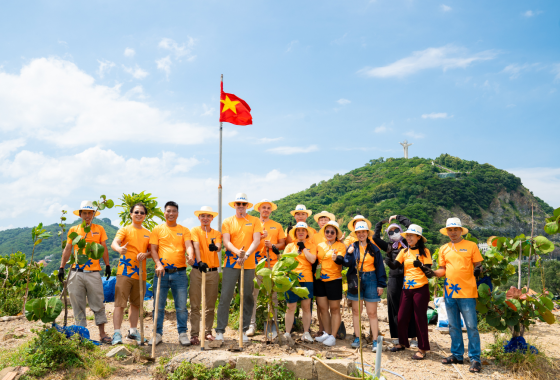 ALPLA team in orange shirts planting trees outdoors, with a Vietnamese flag and green hills behind.